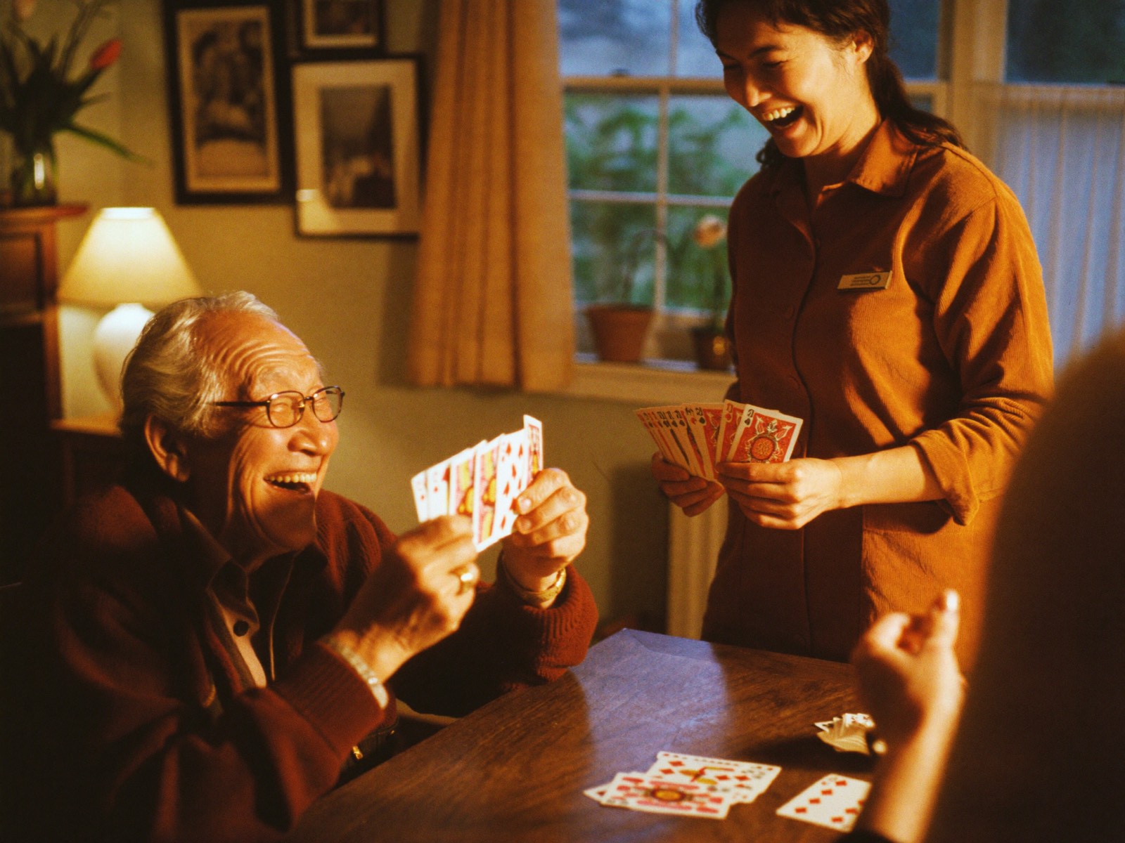 An elderly man laughing heartily during a card game with his caregiver at the dining table