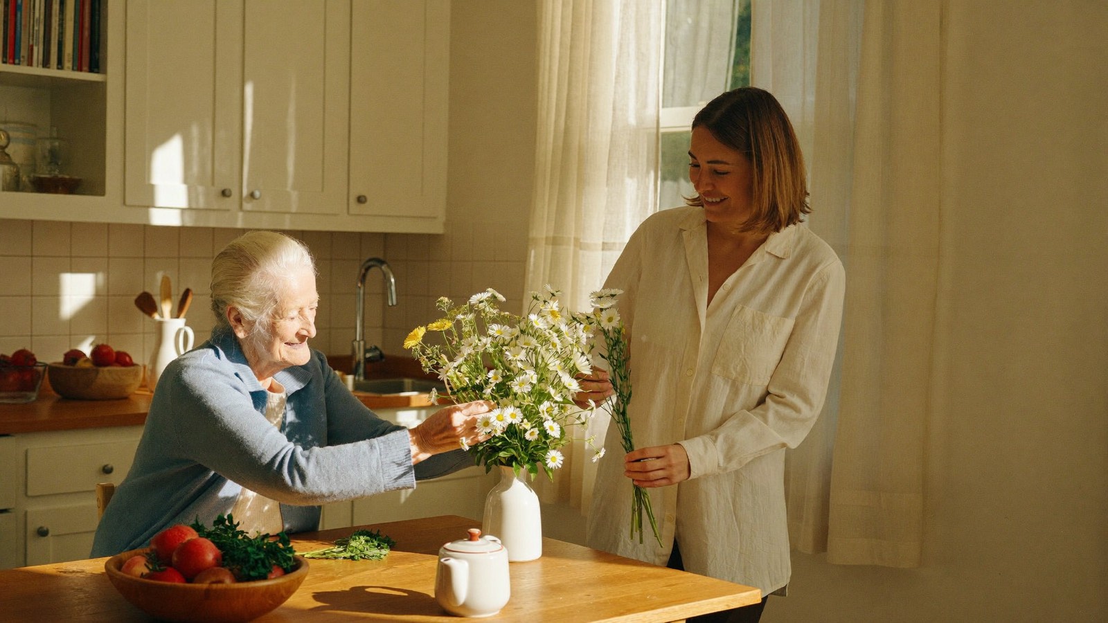 An elderly woman arranging fresh flowers with her caregiver in a sunlit kitchen