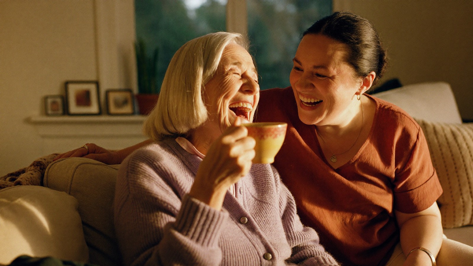 An elderly woman and her caregiver laughing together over tea in warm afternoon light