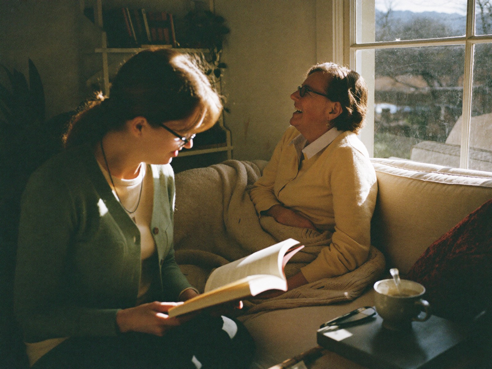 A caregiver reading aloud on the sofa while an elderly woman listens, smiling